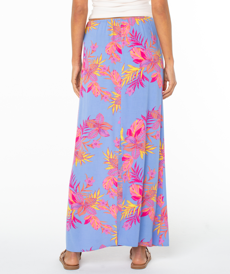 A person is seen from the back wearing the Roxy Iyla Skirt—a long, light blue skirt with pink, orange, and yellow tropical leaves—paired with a white top and brown sandals, set against a white background.