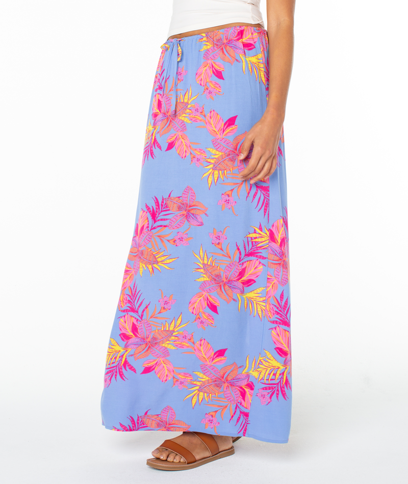A woman wears the Roxy Iyla Skirt, a light blue summer skirt with a vibrant pink, orange, and yellow floral pattern, styled with a white top and brown sandals. Only her lower half is shown.