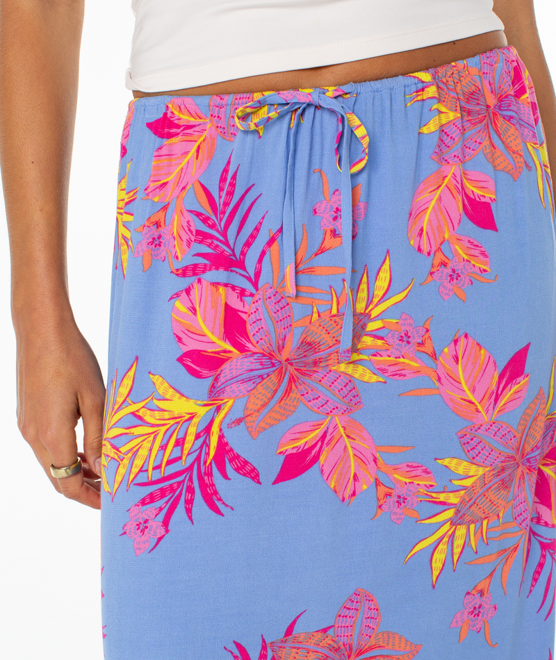 A person wears the Roxy Iyla Skirt, a blue summer skirt with a vibrant tropical floral print in pink, orange, and yellow, paired with a white top. Only their lower torso and part of one arm are visible.