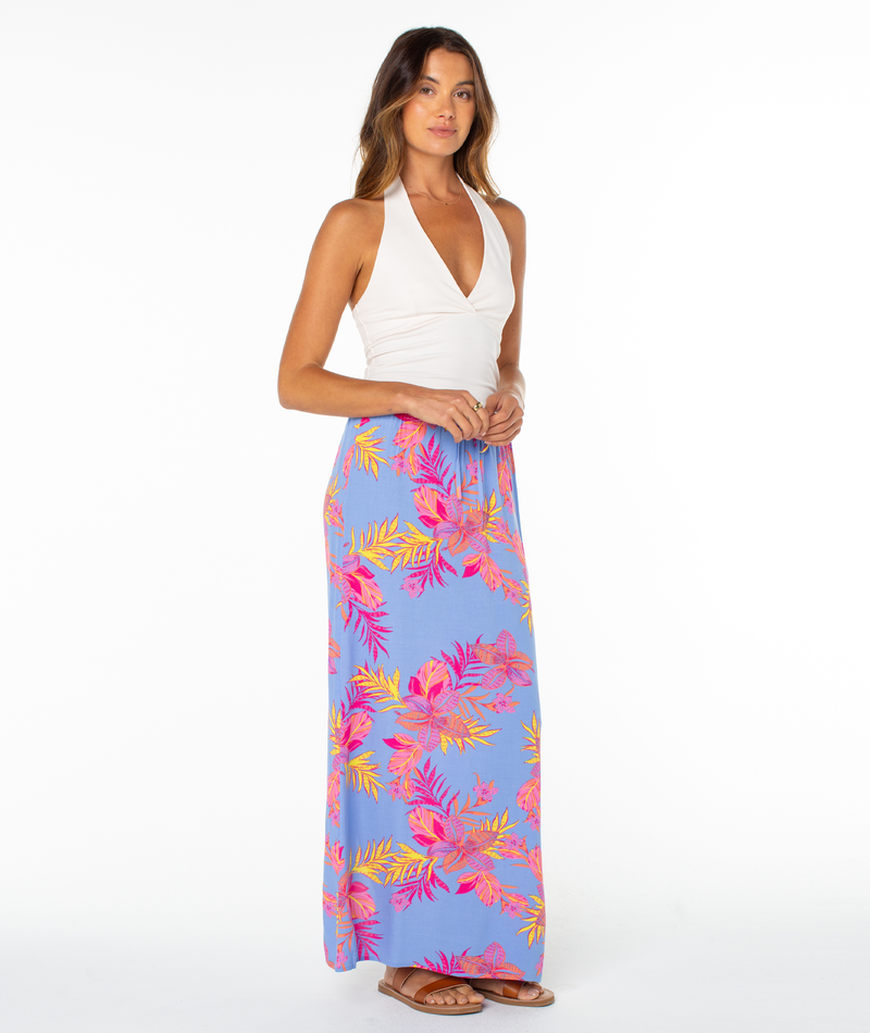 A woman faces forward in a white halter top and the Roxy Iyla Skirt—a long blue summer skirt with vibrant pink, yellow, and orange tropical leaves. She wears sandals against a plain white background.