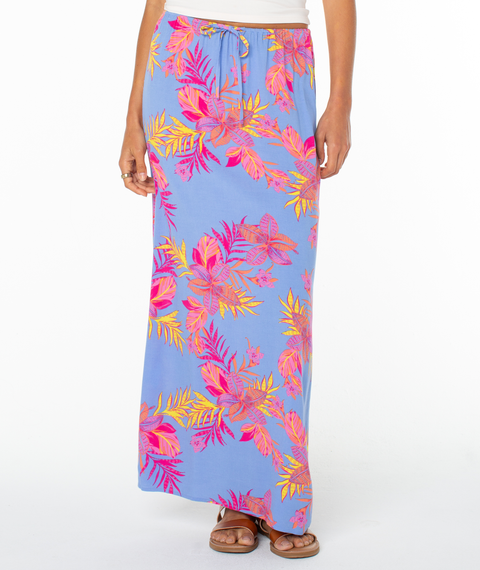 A person wears the Roxy Iyla Skirt, a blue summer skirt with vibrant pink, orange, and yellow tropical leaf prints, paired with sandals and a white top, standing against a white background.