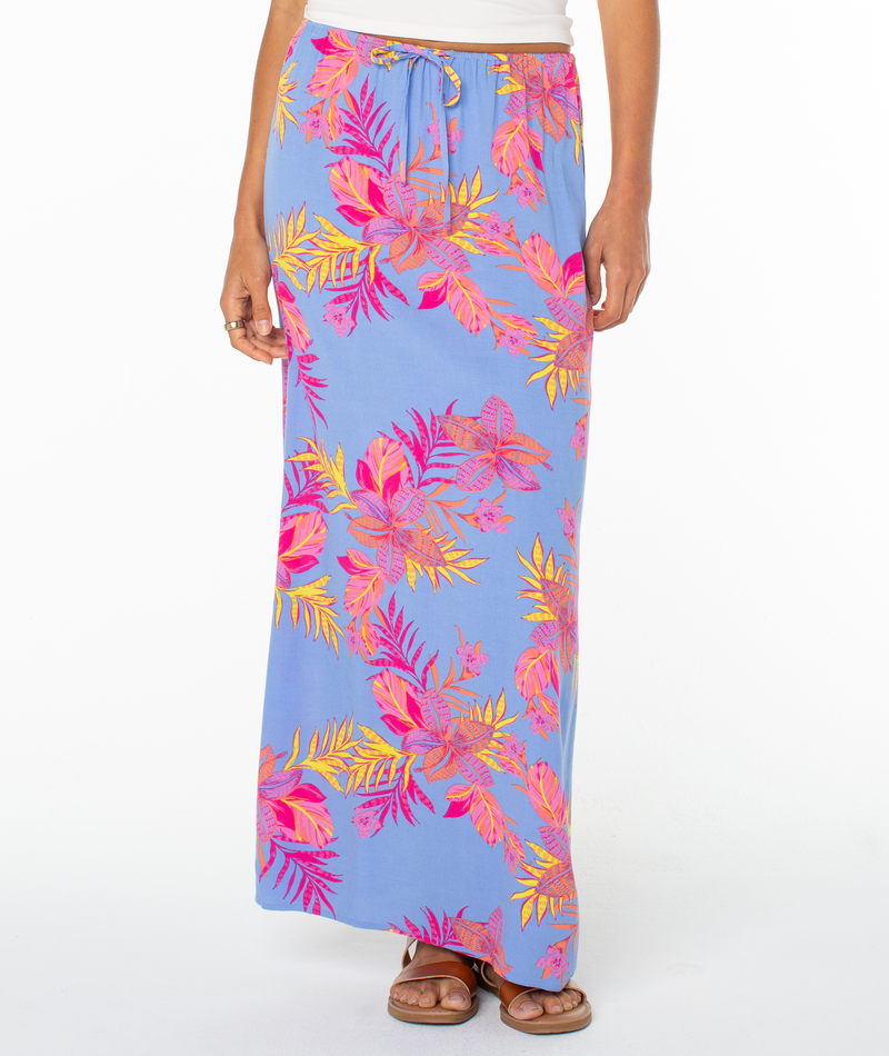 A person wears the Roxy Iyla Skirt, a blue summer skirt with vibrant pink, orange, and yellow tropical leaf prints, paired with sandals and a white top, standing against a white background.