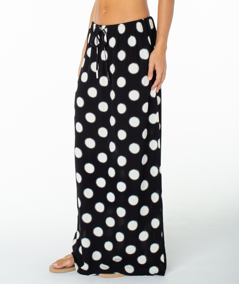 A person wearing the Roxy Iyla Skirt—a black skirt with large white polka dots and a chic tie waist—paired with flat sandals, stands against a white background.