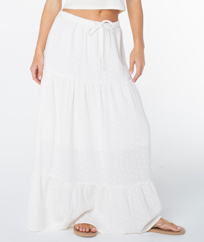 A woman models the Roxy Side Street Skirt, a long white tiered maxi with a subtle dotted pattern and drawstring waist. Only her lower torso, legs, and sandal-clad foot appear against a white background.