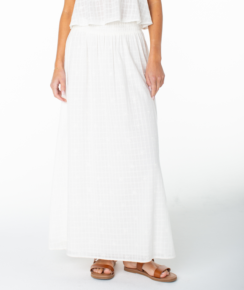 A person wearing a white sleeveless top, the Roxy Sophie Maxi Skirt—a long white cotton piece featuring a subtle checkered pattern—and tan sandals, standing against a plain white background.