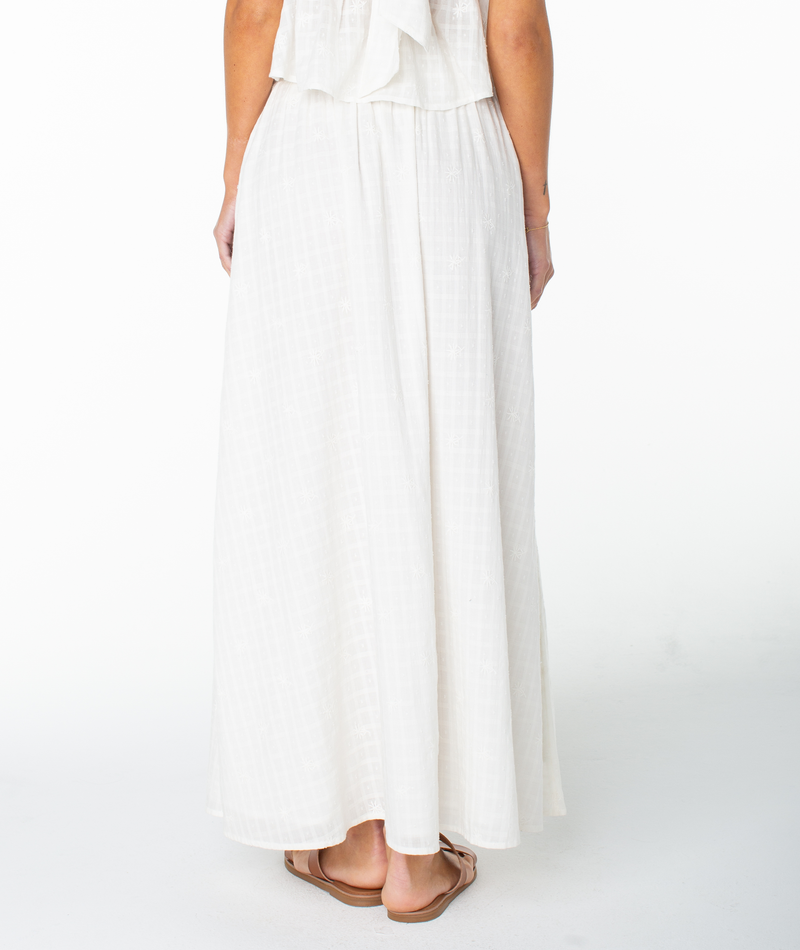 A person stands facing away, wearing the Roxy Sophie Maxi Skirt—a long, white, flowy cotton piece—paired with tan flat sandals. Only the lower half of their body is visible against a plain white background.