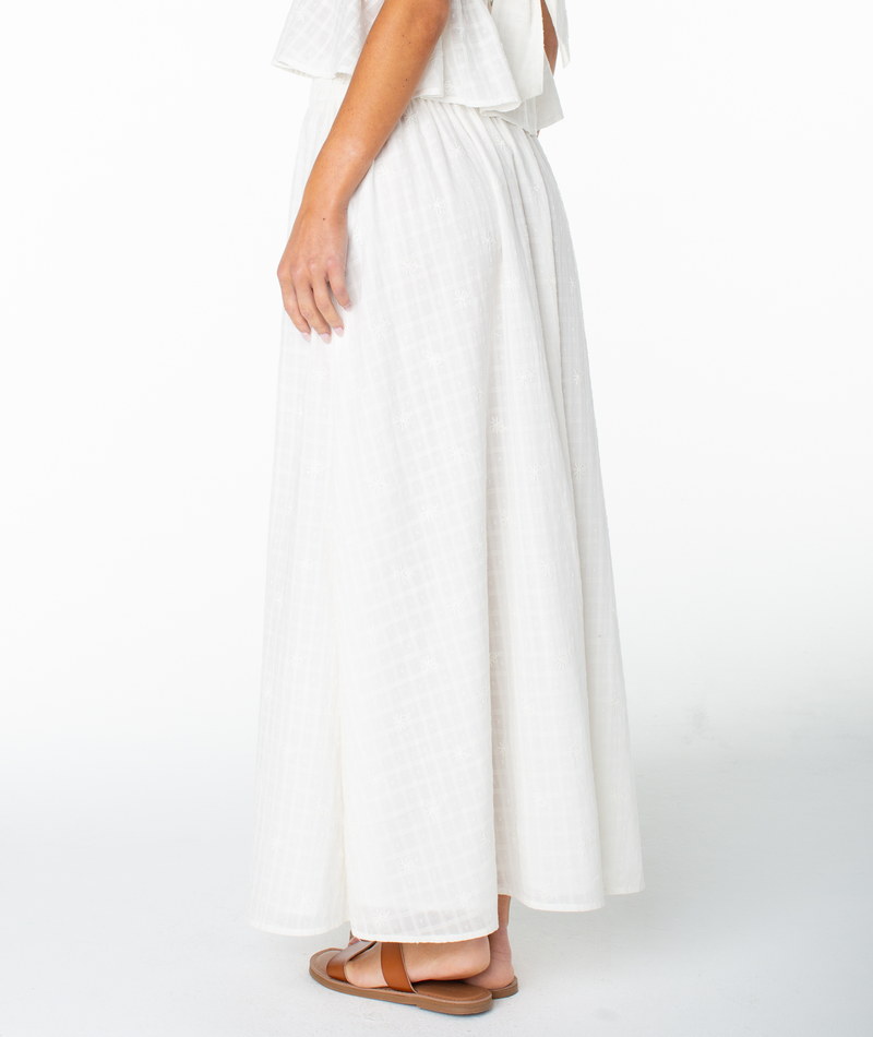 A woman stands sideways against a plain white background wearing the Roxy Sophie Maxi Skirt, a long textured white cotton skirt, paired with brown flat sandals and her left arm relaxed by her side.