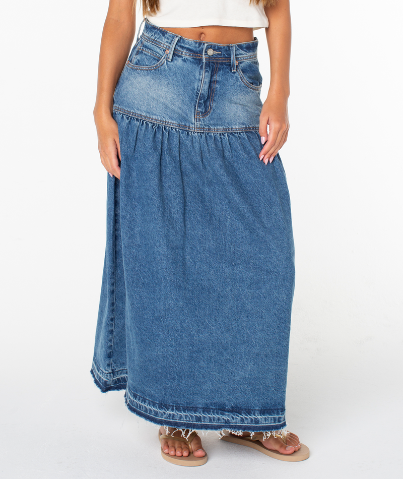 A woman models the Roxy Nocturne Prairie Skirt—a vintage-inspired blue denim maxi with a frayed hem—paired with a white crop top and brown sandals. Her hands rest on the skirt, set against a plain white background.
