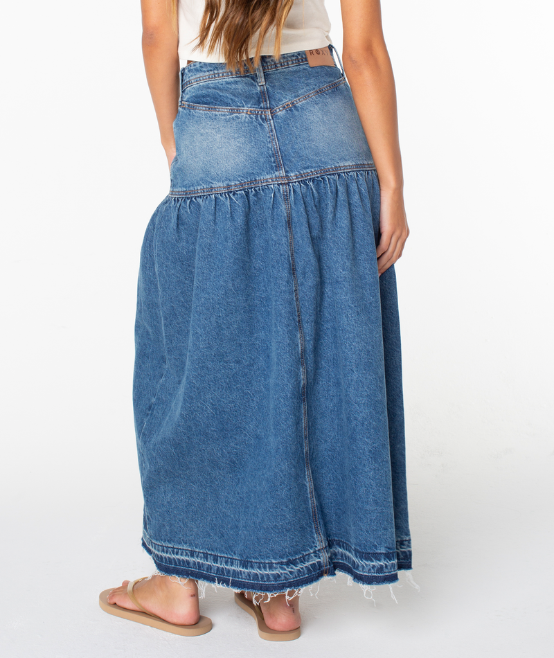 A woman with long brown hair stands against a plain white background, wearing the Roxy Nocturne Prairie Skirt—a vintage-inspired dark wash skirt with a frayed hem—paired with sandals and shown from the back.