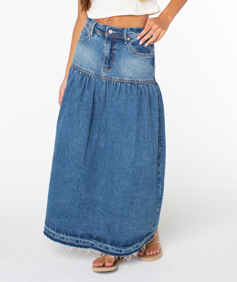 A woman is seen wearing the Roxy Nocturne Prairie Skirt—a long, dark wash denim skirt with a gathered seam at the hip, frayed hem, and visible seams—styled with a white crop top and sandals. Her face is not visible.