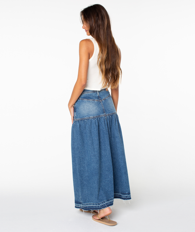 A woman stands sideways against a plain white background, wearing the Roxy Nocturne Prairie Skirt—a long, vintage-inspired denim skirt—paired with a white sleeveless top and sandals. She has long brown hair, hands in her pockets, and is looking down.