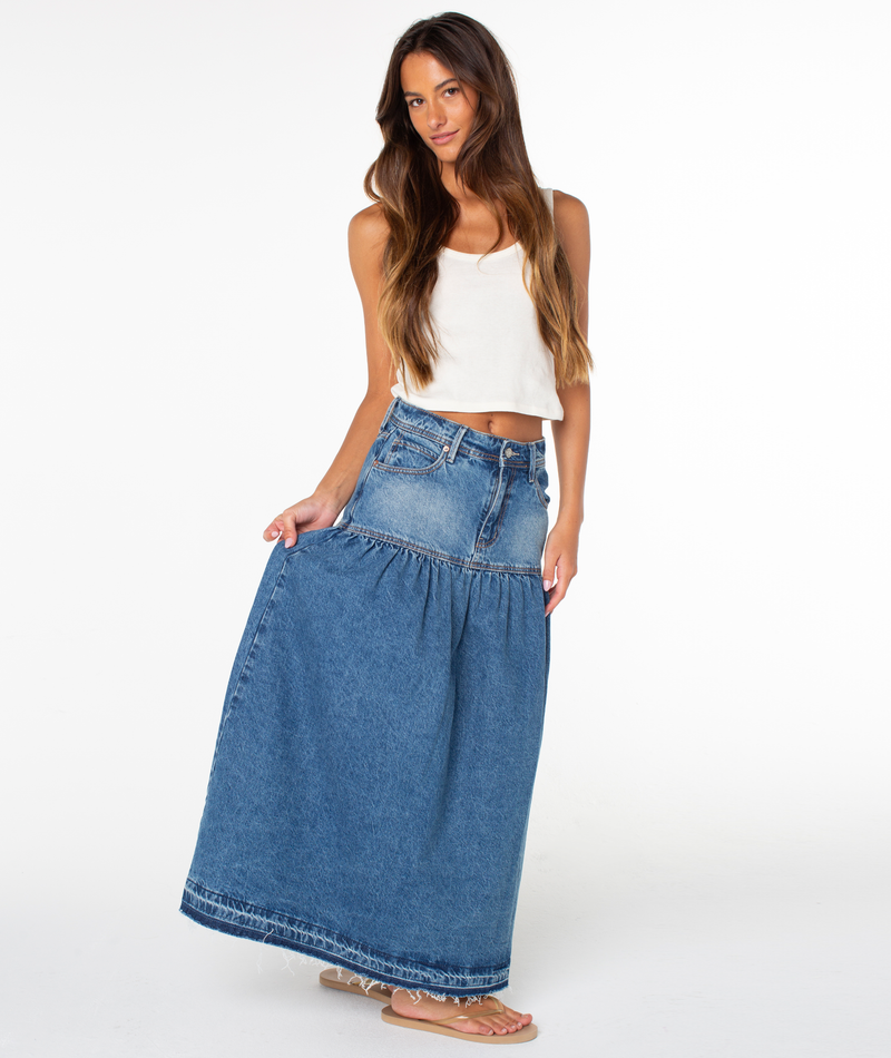 A woman poses against a plain white background, wearing a white tank top, sandals, and the Roxy Nocturne Prairie Skirt in Salt Fade Denim Wash, holding one side of her vintage-inspired skirt slightly out.
