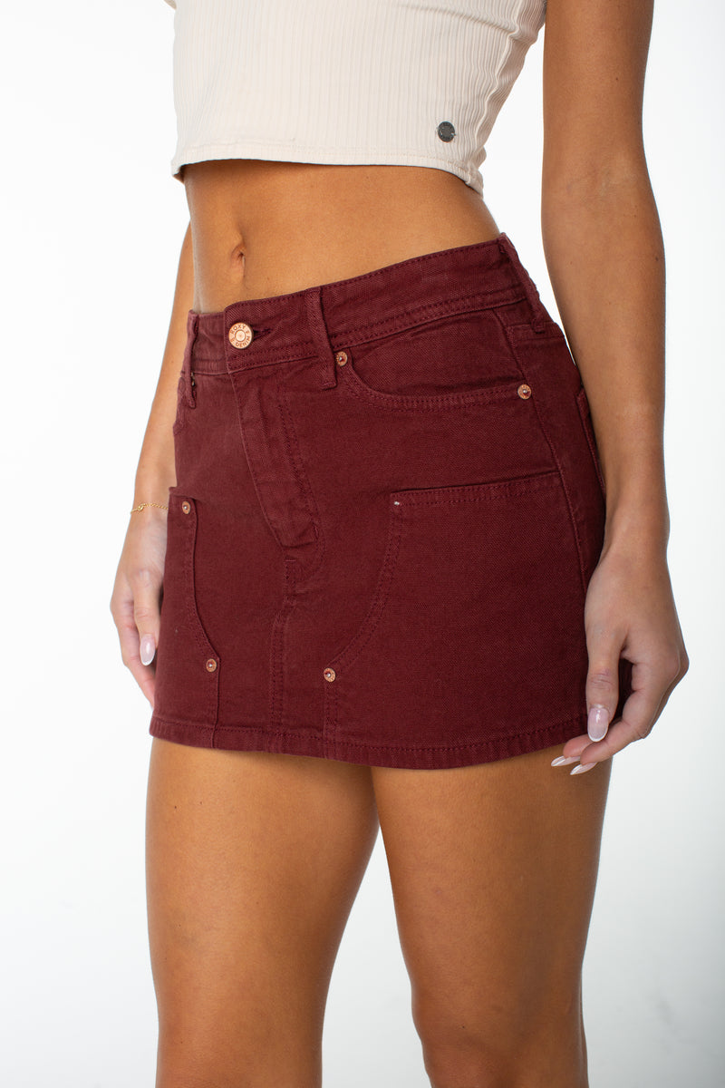 A person models the Roxy Reel Life Skirt—a high-waisted, dark red denim mini skirt with subtle carpenter details—paired with a cream crop top. Shown from the torso to upper legs against a plain white background, the image highlights a distinctly feminine silhouette.