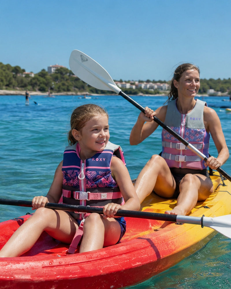 A woman and a girl, with the girl wearing the Roxy Child CGA Vest by Roxy, paddle a yellow and red kayak on clear blue water under a sunny sky. Trees and buildings are visible in the background, highlighting the importance of water safety during their adventure.
