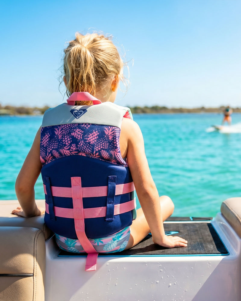A young child wearing the Roxy Child CGA Vest by Roxy sits on the edge of a boat, looking out at the bright blue water on a sunny day, ensuring both comfort and water safety.