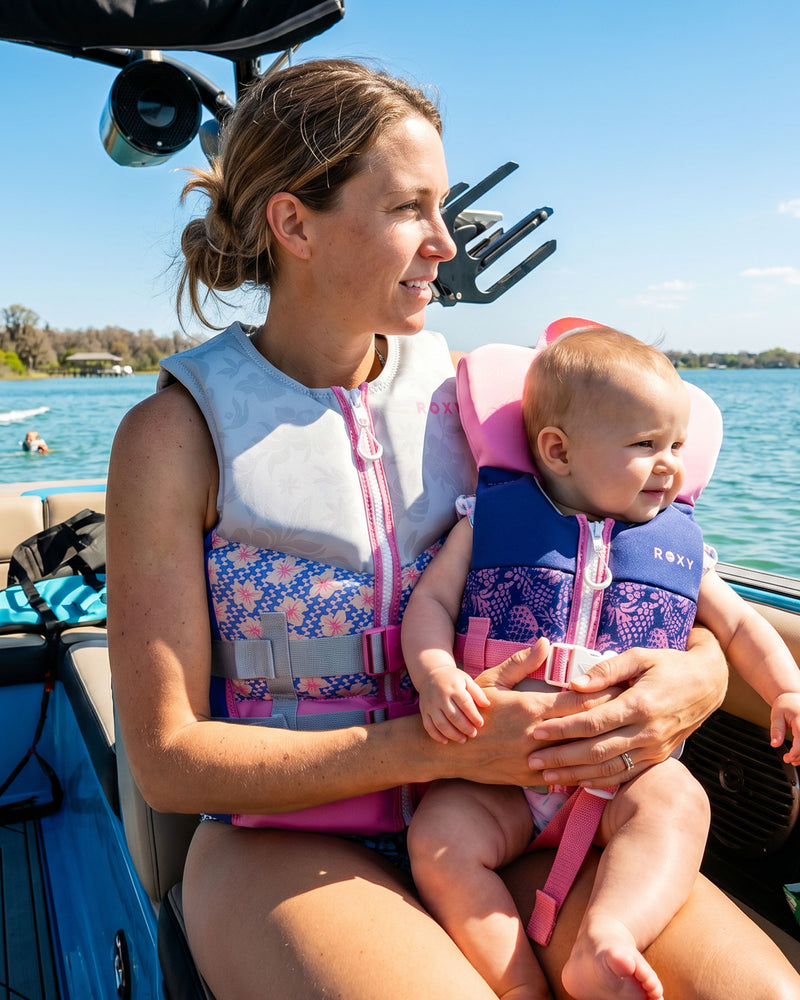 A woman holds a smiling baby wearing the Roxy Infant CGA Vest, a USCG approved life jacket by Roxy, as they sit together on a boat and look out over the water on a sunny day.