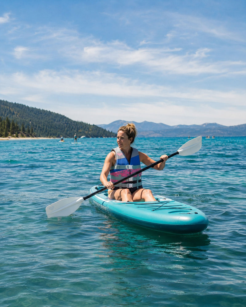 A woman paddles a teal kayak on a clear blue lake, wearing the Roxy Womens Duraweave Vest by Roxy. Forested hills and mountains are visible in the background under a partly cloudy sky.