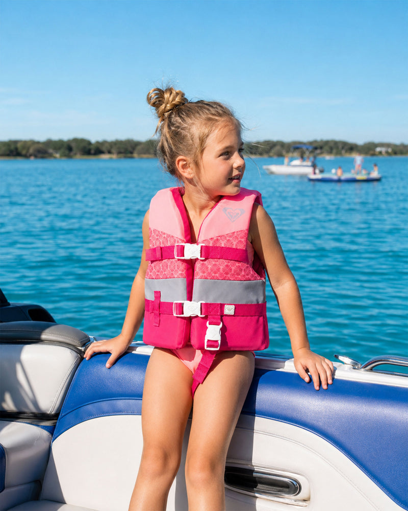 A young girl wearing a Roxy Child Duraweave Vest sits on the edge of a boat, smiling and looking to the side. The boat is on bright blue water, with people and trees visible in the background under a clear sky.