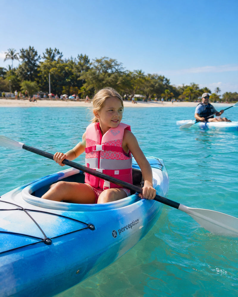 Wearing a Roxy Child Duraweave Vest, a young girl paddles a blue kayak across clear turquoise water, with another kayaker and a sandy beach fringed with trees in the background beneath a bright blue sky.