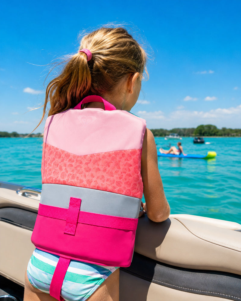 A young girl wearing a Roxy Child Duraweave Vest by Roxy stands on a boat, gazing out over bright blue water toward inflatable rafts with people in the distance on a sunny day.