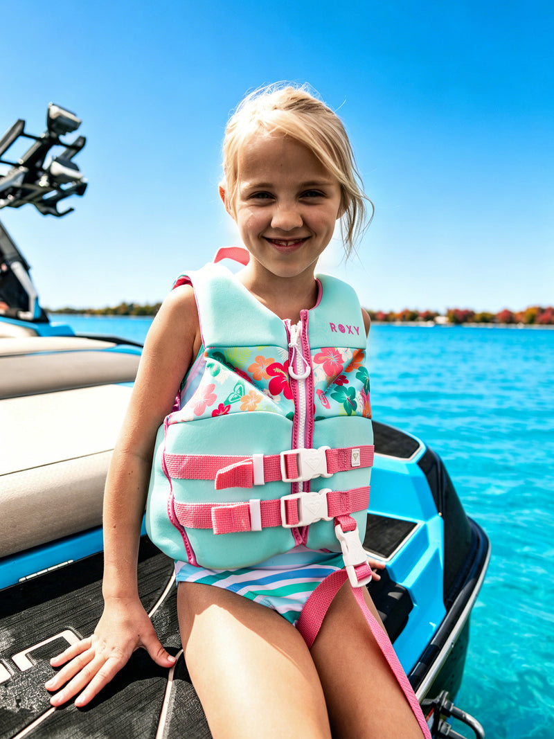 A smiling girl wearing a Roxy Child Daily CGA Vest by Roxy sits on a boat in bright sunlight, with clear blue water and a distant treeline in the background.