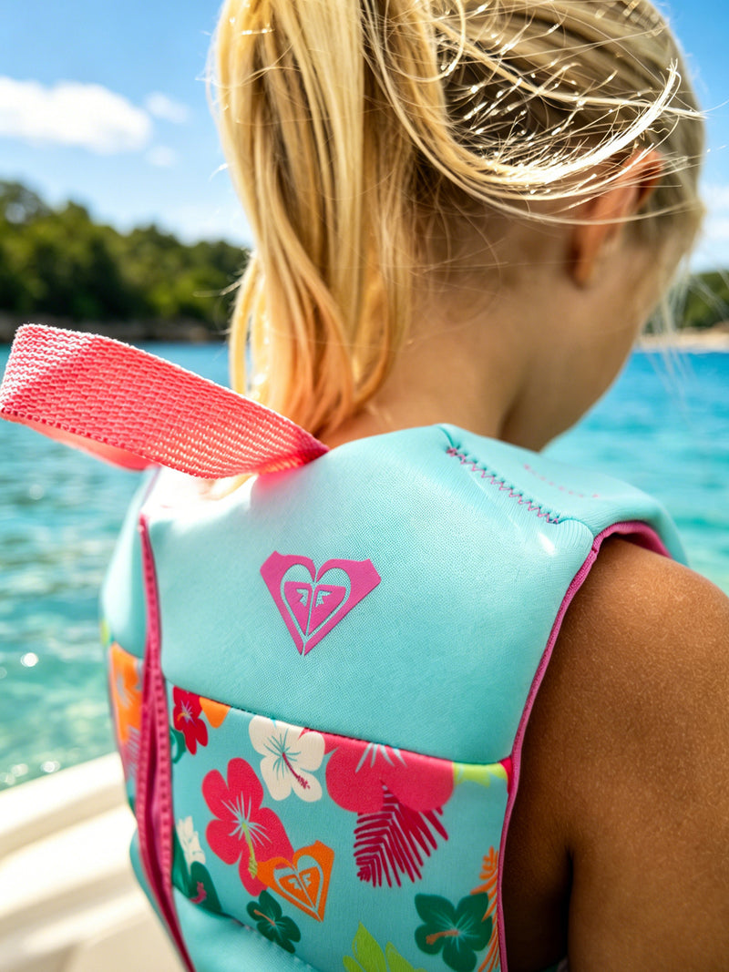 A young girl with blonde hair in a ponytail wears the Roxy Child Daily CGA Vest by Roxy, a USCG-approved kids’ life jacket featuring a vibrant floral pattern and heart logo, as she stands near clear blue water on a sunny day.