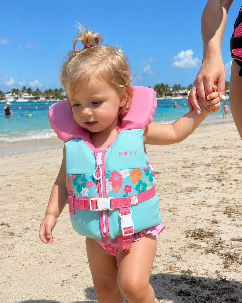 A toddler wearing a pink and turquoise floral Roxy Infant Daily CGA Vest walks along a sandy beach, holding an adult’s hand. The USCG approved Roxy life vest ensures the child’s safety, while the ocean and swimmers can be seen in the background beneath a bright blue sky.