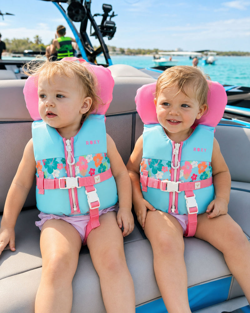 Two young children wearing Roxy Infant Daily CGA Vests are seated side by side on a boat. Both have light brown hair and are smiling while looking in different directions. Their USCG-approved infant life vests from the brand Roxy feature a cheerful floral pattern with pink trim. The background includes water and other boats.