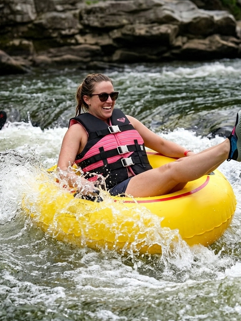 A woman wearing Roxy Women’s Oasis Duraweave sunglasses laughs while riding a yellow inner tube through splashing river rapids, with rocks in the background.