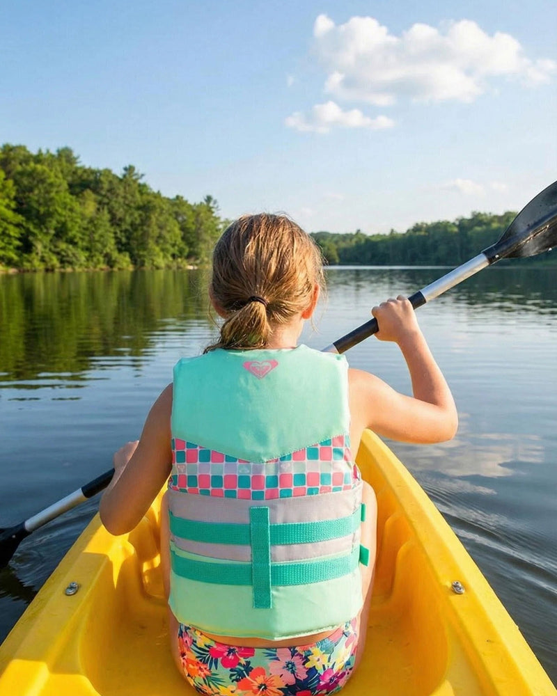 Wearing a vibrant Roxy Youth Oasis CGA Vest by Roxy, a child paddles a yellow kayak across a calm lake encircled by trees beneath a blue sky dotted with clouds, enjoying a safe day of boating.