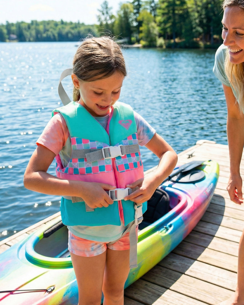 A young girl wearing a vibrant Roxy Child Oasis CGA Vest, a USCG-approved life vest by Roxy designed for children’s water safety, stands on a wooden dock next to a kayak, smiling as she fastens her buckle. An adult woman stands nearby, with trees and a lake visible in the background.