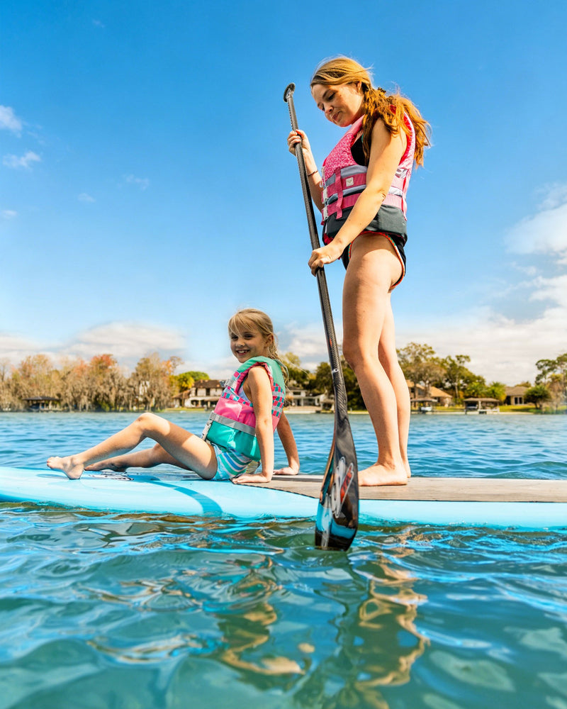 Two girls wearing Roxy Child Oasis CGA Vests paddleboard on a lake under a bright blue sky. Practicing kids water safety in their Roxy life vests, one girl stands with a paddle while the other sits and smiles. Trees and houses are visible in the background.