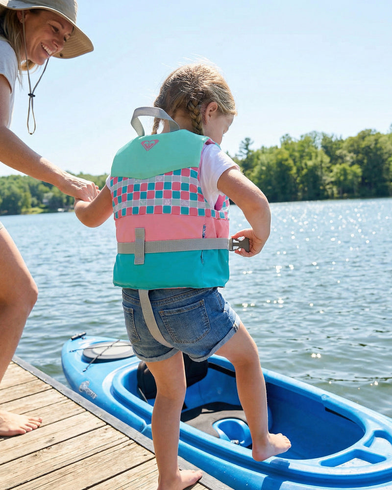 A woman assists a young girl wearing a Roxy Child Oasis CGA Vest by Roxy as she steps into a blue kayak from a wooden dock on a sunny lakeside day. Trees and blue sky in the background highlight the importance of kids’ water safety.