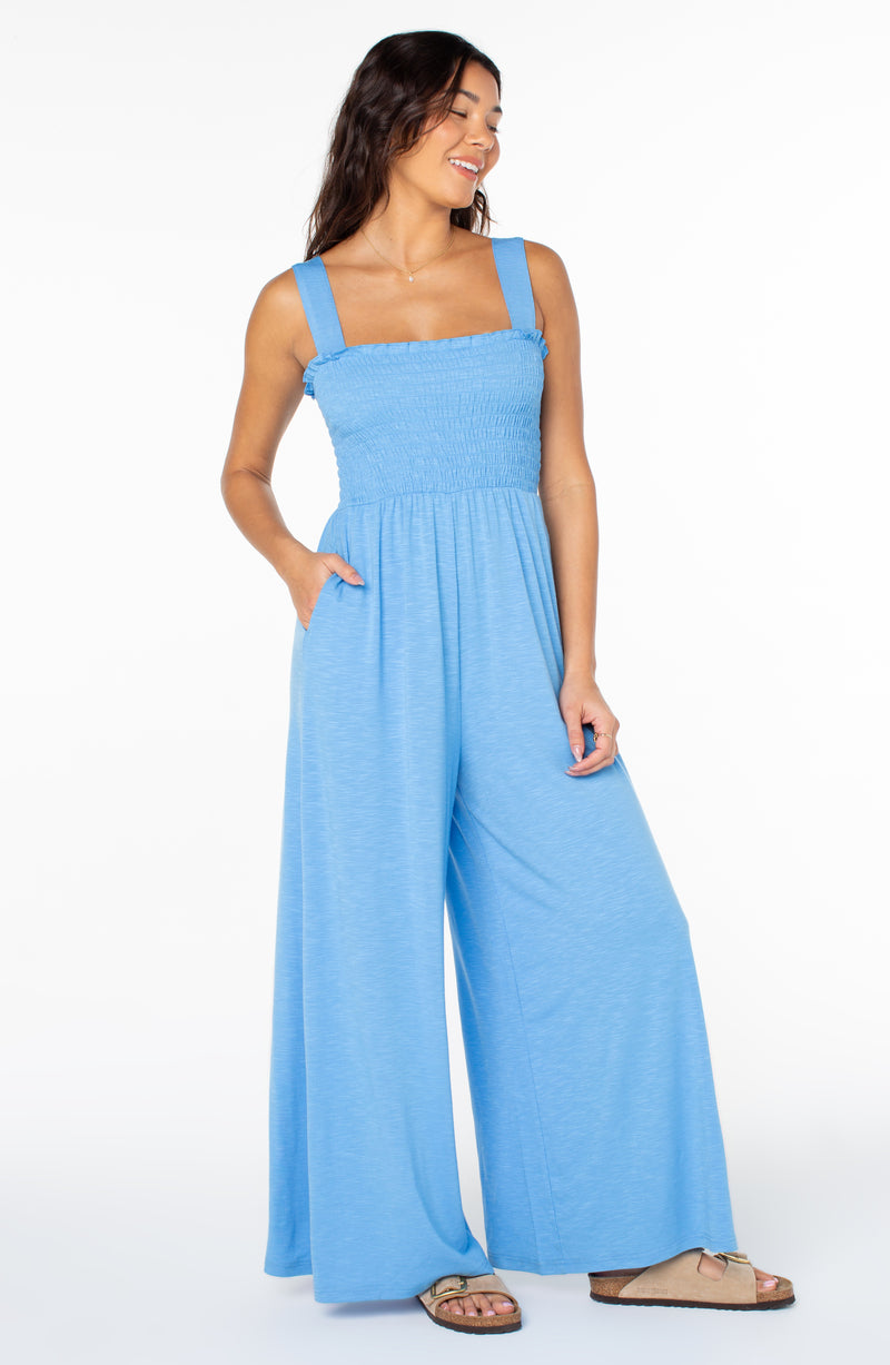 A person wears a light blue Roxy Just Passing By Jumpsuit featuring a sleeveless smocked bodice and wide-leg trousers.