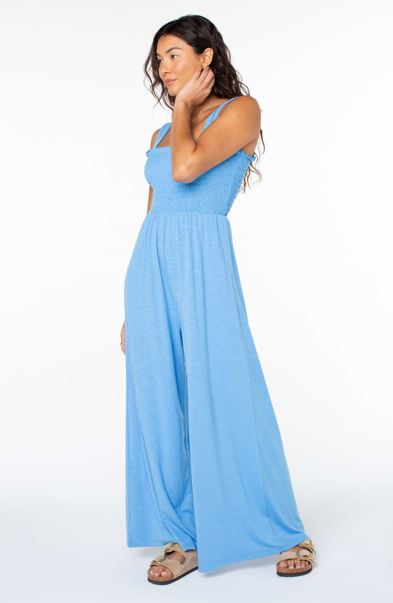 A woman wearing a light blue, sleeveless Just Passing By Jumpsuit featuring a smocked bodice and wide-leg trousers.