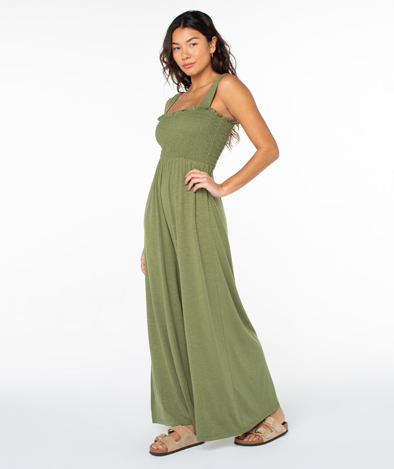 A person wearing the olive green Roxy Just Passing By Jumpsuit with a smocked bodice and wide-leg trousers, paired with tan slide sandals.