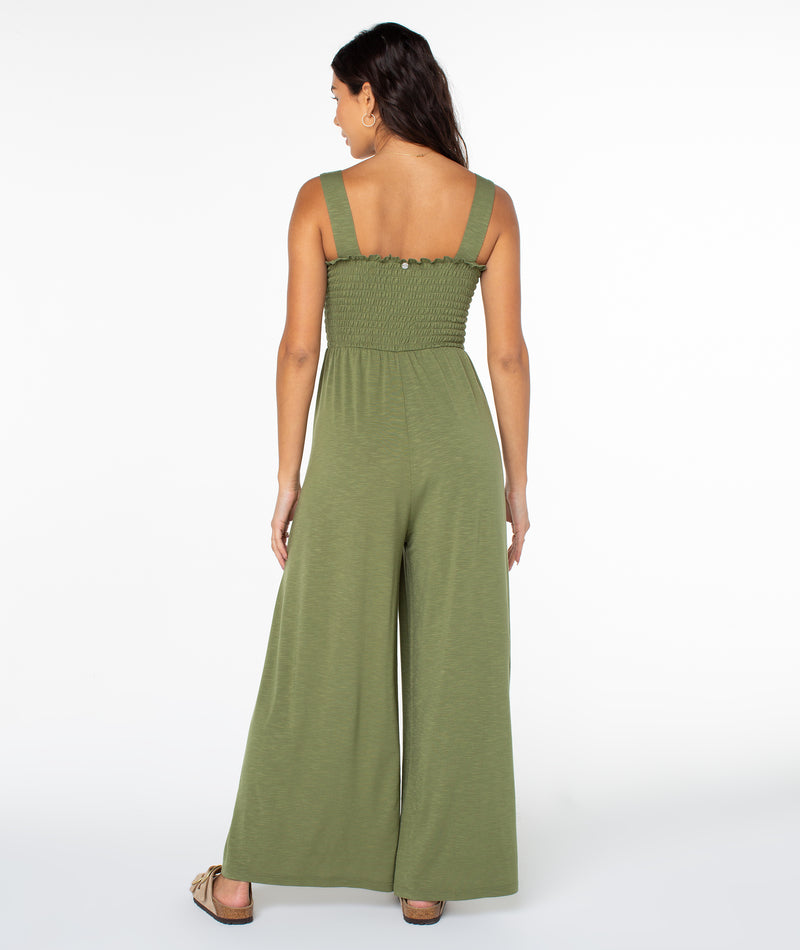 Back view of a woman wearing the olive green Roxy Just Passing By Jumpsuit, featuring a sleeveless smocked bodice and wide-leg trousers.