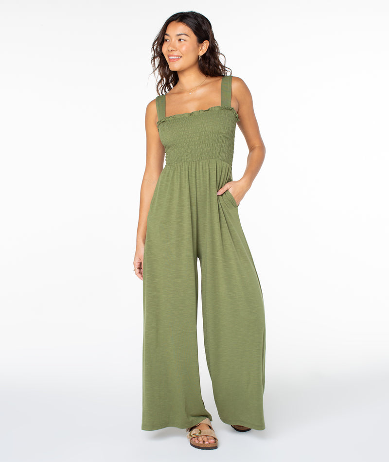 A person wearing a Roxy Just Passing By Jumpsuit in olive green, featuring a sleeveless smocked bodice and wide-leg trousers.