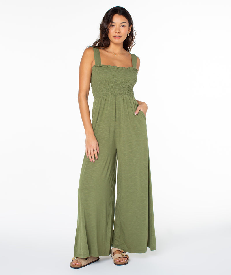A person wearing a loden green Roxy Just Passing By Jumpsuit with a square, smocked bodice and wide-leg pants.