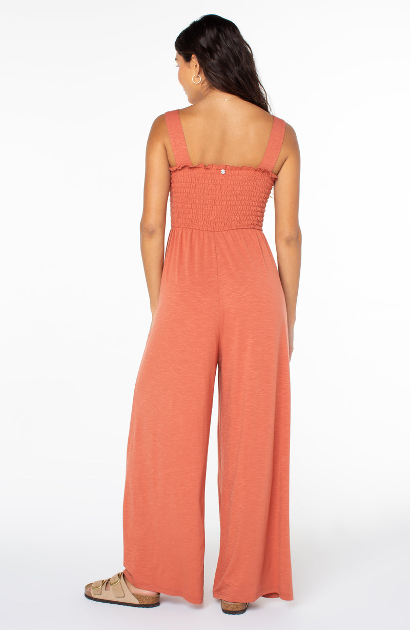 Back view of a person wearing a rust-colored, sleeveless Just Passing By Jumpsuit with a smocked bodice and wide-leg trousers, paired with tan slide sandals.