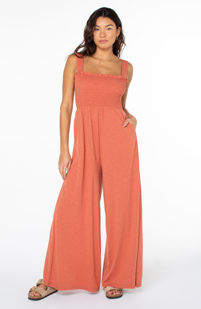 A person wearing a burnt orange, sleeveless, wide-leg Roxy Just Passing By Jumpsuit with a smocked bodice and sandals.
