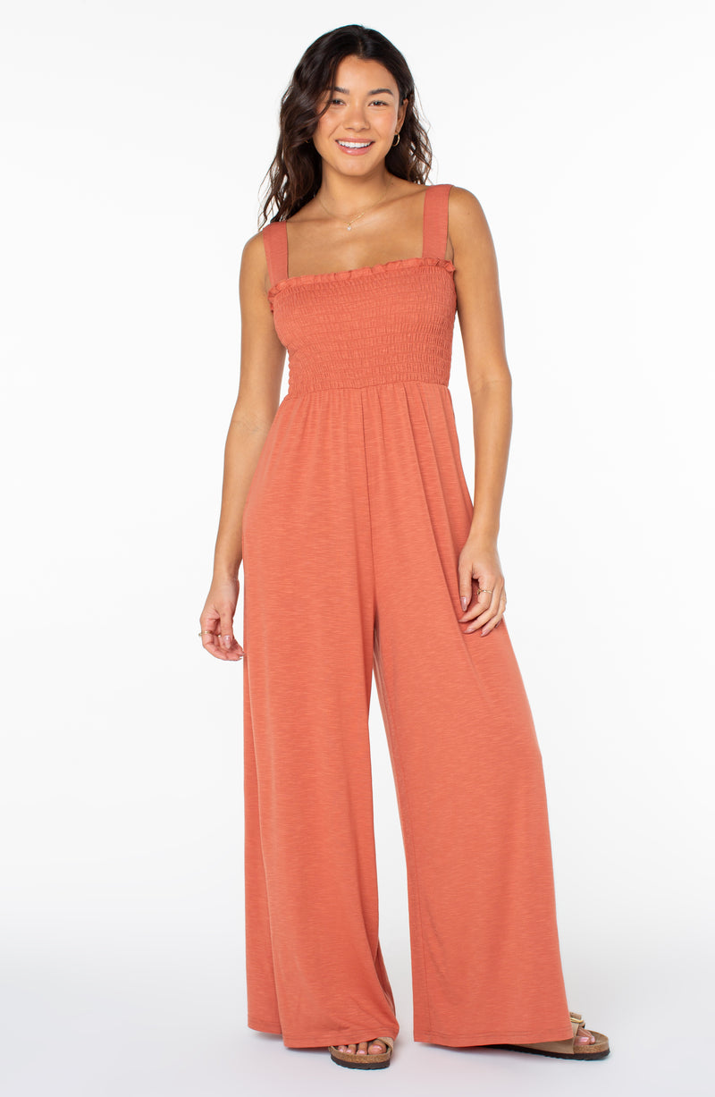 A person wears a Roxy Just Passing By Jumpsuit in Aragon, featuring a burnt-orange smocked bodice, wide-leg trousers, and thick shoulder straps.