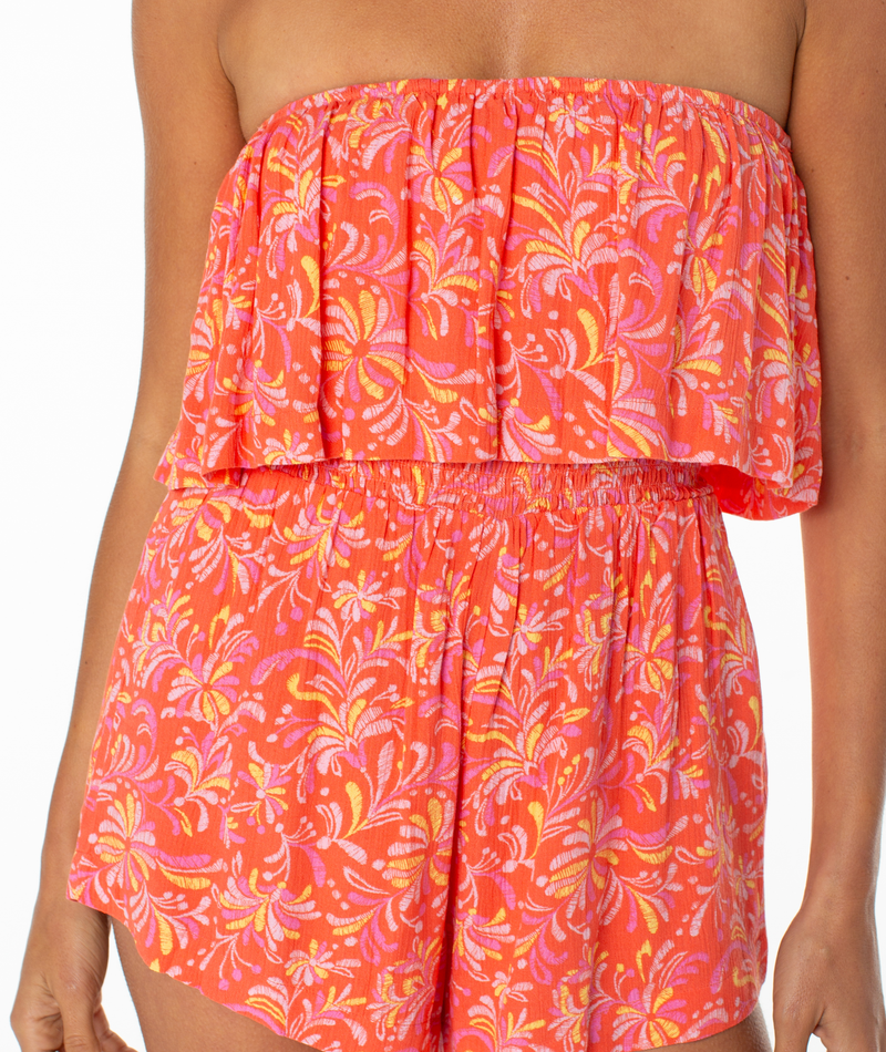 A woman wears the Roxy Lets Go Romper, a coral strapless outfit with a ruffled top and yellow-pink leafy print. The flowy shorts create a breezy look, showing her torso and upper legs.