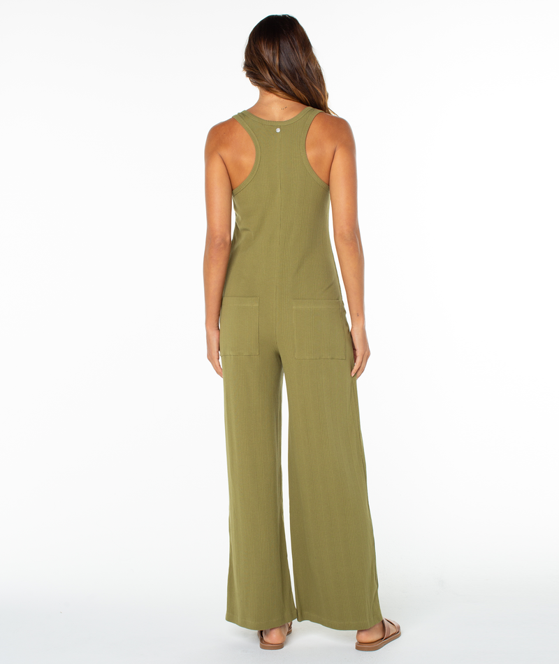 A woman with long hair wears the olive green Roxy Running Tide Jumpsuit, featuring wide legs and large back pockets, channeling a ’70s vibe. She stands in sandals facing away against a plain white background.