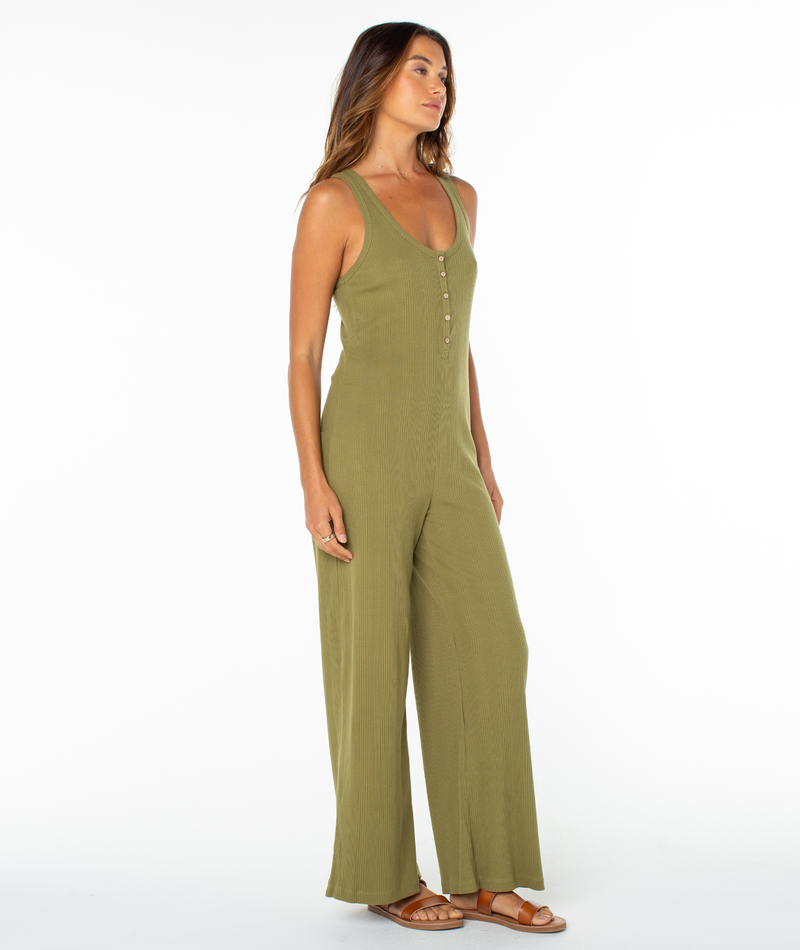 A woman with long hair and bare feet wears the Roxy Running Tide Jumpsuit, a sleeveless olive green style with front buttons and wide-leg pants, posing against a plain white background with subtle 70’s vibes.