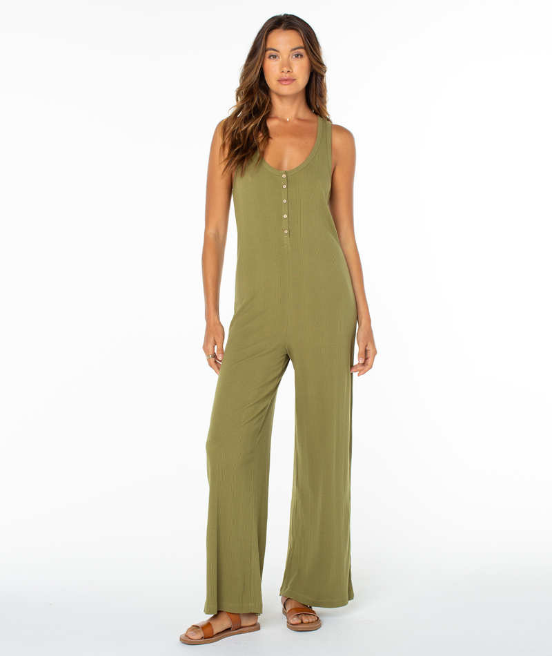 A woman faces forward in the Roxy Running Tide Jumpsuit, a sleeveless olive green piece with wide legs and button details. Her long wavy hair and brown sandals complete the look against a plain white background, evoking subtle ’70s style.
