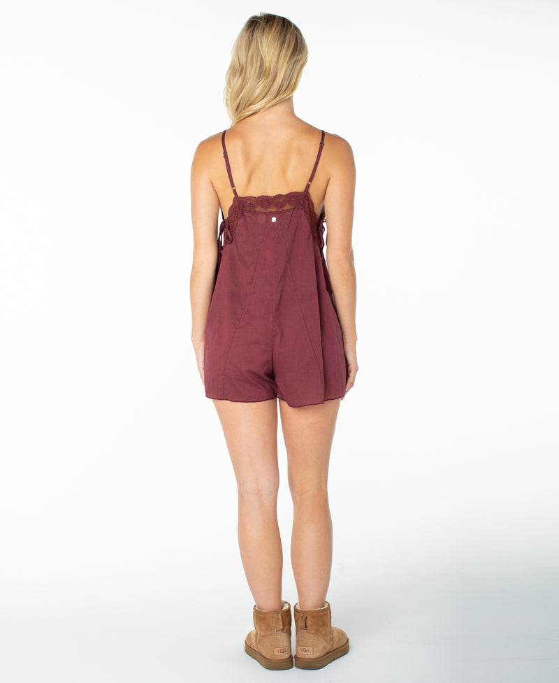 A woman stands facing away, wearing the Roxy Moonrise Romper—a sleeveless, maroon cotton-blend one-piece with lace details—paired with tan ankle boots. Her blond hair falls loosely, set against a plain white background that emphasizes this versatile all-in-one outfit and true wardrobe staple.