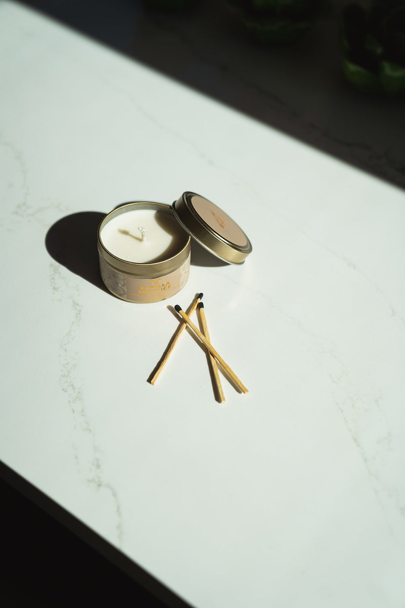 An open Vanilla Solstice Holiday Tin Candle in a gold metal tin sitting next to three wooden matches.