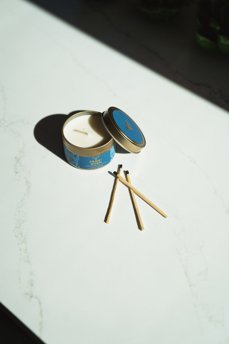 An open ROXY Frost Rush Holiday Tin Candle with a blue label and its lid resting beside it, accompanied by three wooden matches on a white surface.