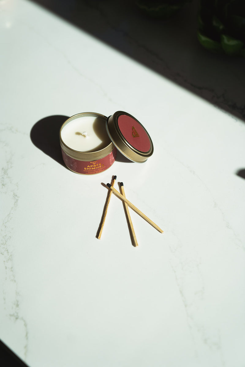 An open, pink-labeled Apres Shimmer Holiday Tin Candle with its lid resting beside it, accompanied by three matches on a white surface.